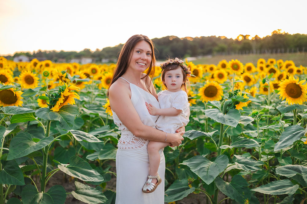 maman qui porte son enfant qui porte une couronne de fleurs devant tournesol plan americain lisa mazeau photographe professionnelle gironde